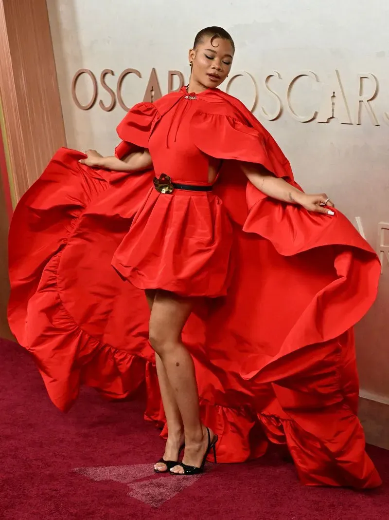 نجمة ترتدي فستانًا أحمرًا مزينًا بأكمام كبيرة، تتألق على السجادة الحمراء لحفل الأوسكار، مع خلفية تحمل شعار "أوسكار".