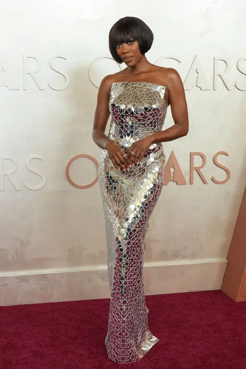 إطلالة مميزة لنجمة ترتدي فستاناً فضياً عاكساً للضوء على السجادة الحمراء في حفل الأوسكار، مع خلفية تحمل شعار الأوسكار.