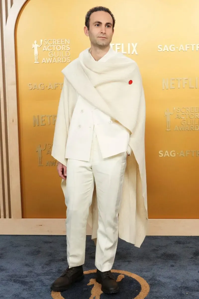 رجل يرتدي بدلة بيضاء مع عباءة بيضاء وحذاء داكن، يقف على السجادة الحمراء في حفل جوائز نقابة ممثلي الشاشة.