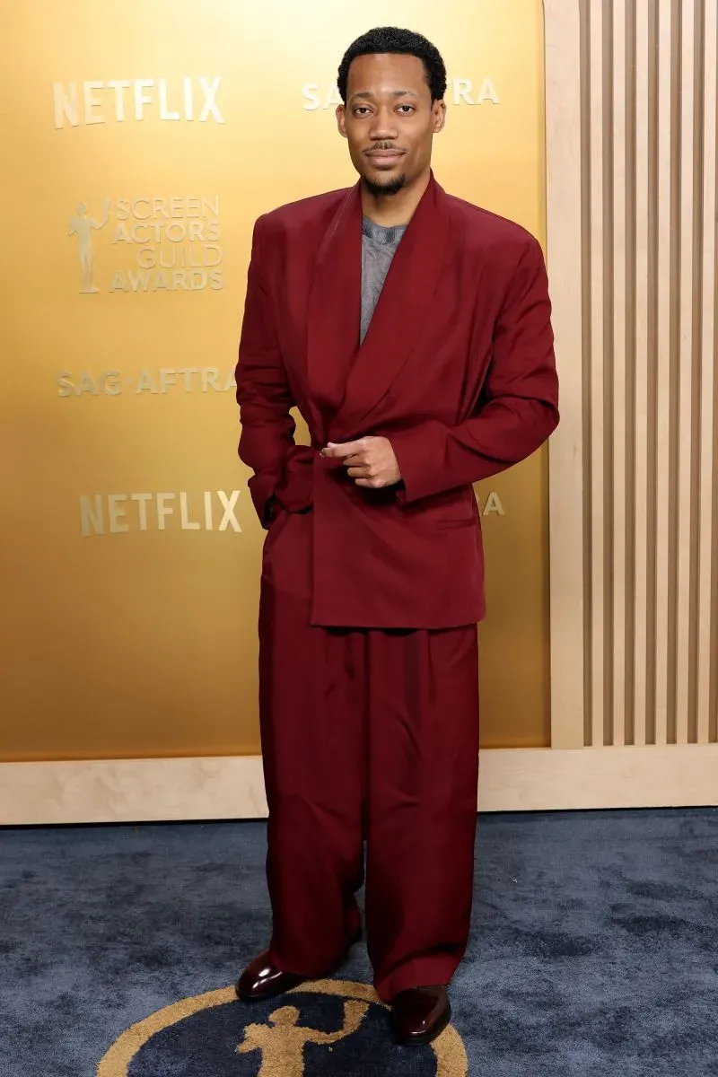 رجل يرتدي بدلة حمراء أنيقة، يقف على السجادة الحمراء لحفل توزيع جوائز نقابة الممثلين، مع خلفية ذهبية تحمل شعار الحدث.