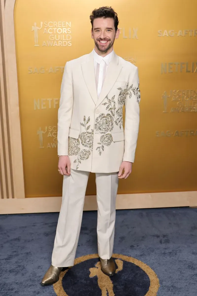 رجل يرتدي بدلة بيضاء مزينة بتطريزات زهرية، يقف مبتسمًا على السجادة الحمراء لحفل جوائز نقابة ممثلي الشاشة.