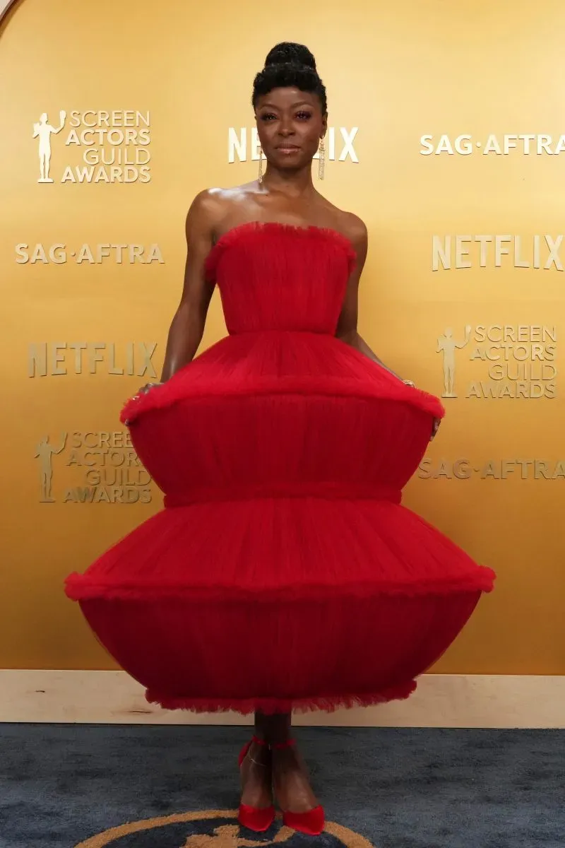 امرأة ترتدي فستاناً أحمر جريئاً ذو تصميم فريد يشبه الكعكة، تقف أمام خلفية حفل جوائز نقابة ممثلي الشاشة.