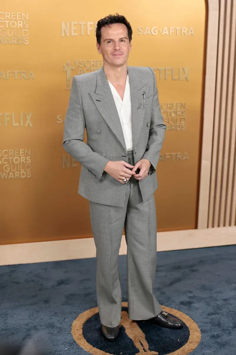 رجل يرتدي بدلة مربعة رمادية اللون مع قميص أبيض، يقف مبتسمًا أمام خلفية حفل جوائز نقابة ممثلي الشاشة.