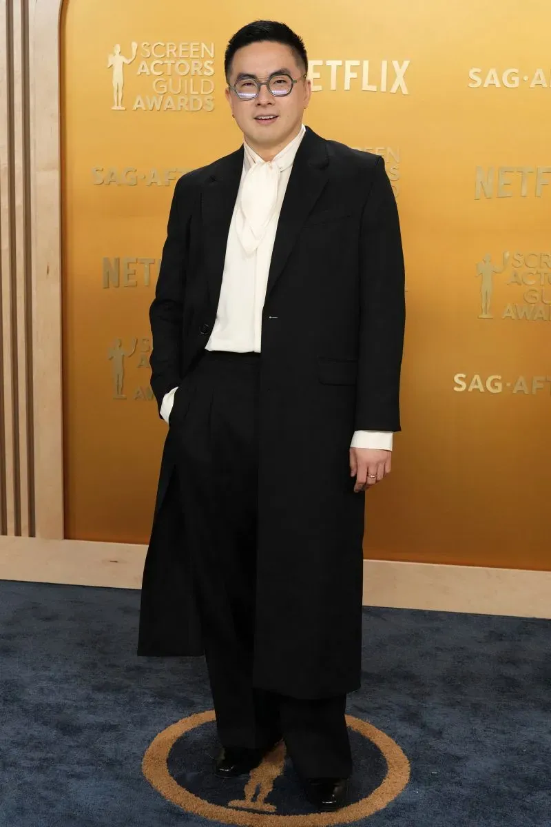 شخص يرتدي بدلة سوداء أنيقة وقميص أبيض، يقف على السجادة الحمراء خلال حفل توزيع جوائز نقابة ممثلي الشاشة مع خلفية ذهبية.