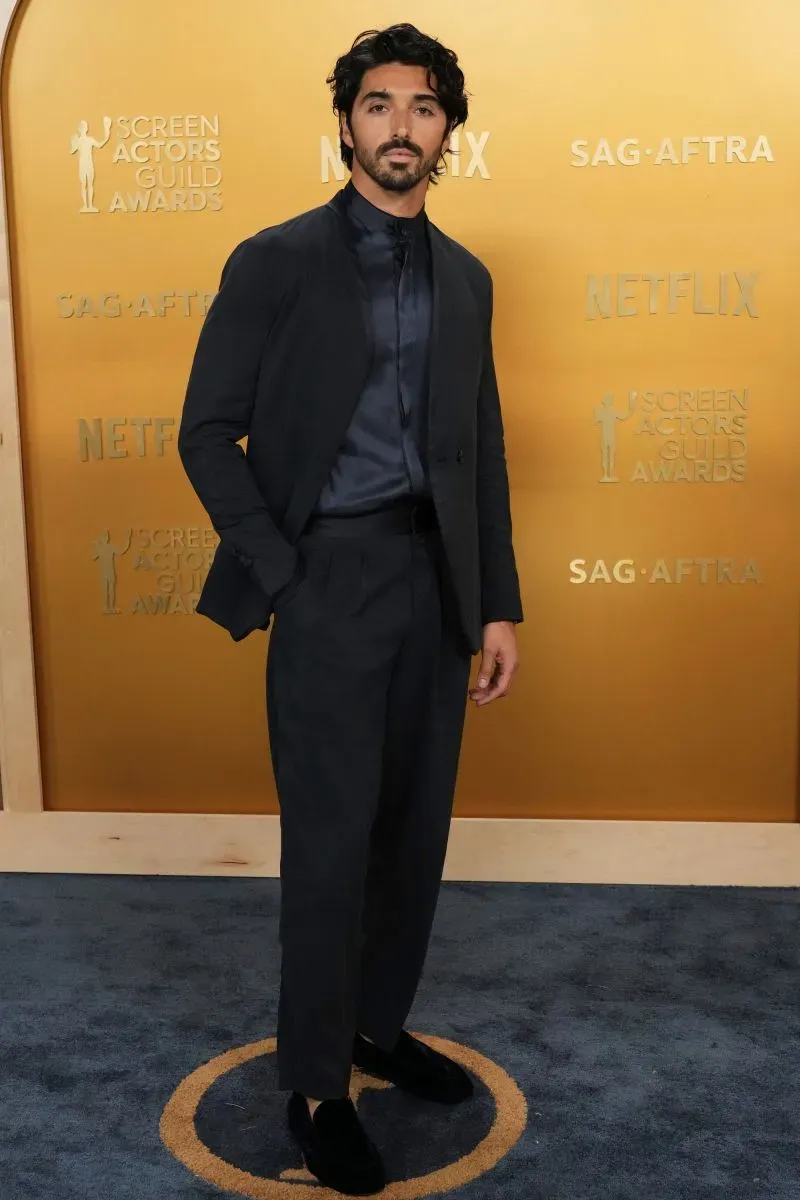 رجل يرتدي بدلة داكنة أنيقة يقف على السجادة الحمراء لحفل جوائز نقابة ممثلي الشاشة، مع خلفية ذهبية تحمل شعارات الحفل.