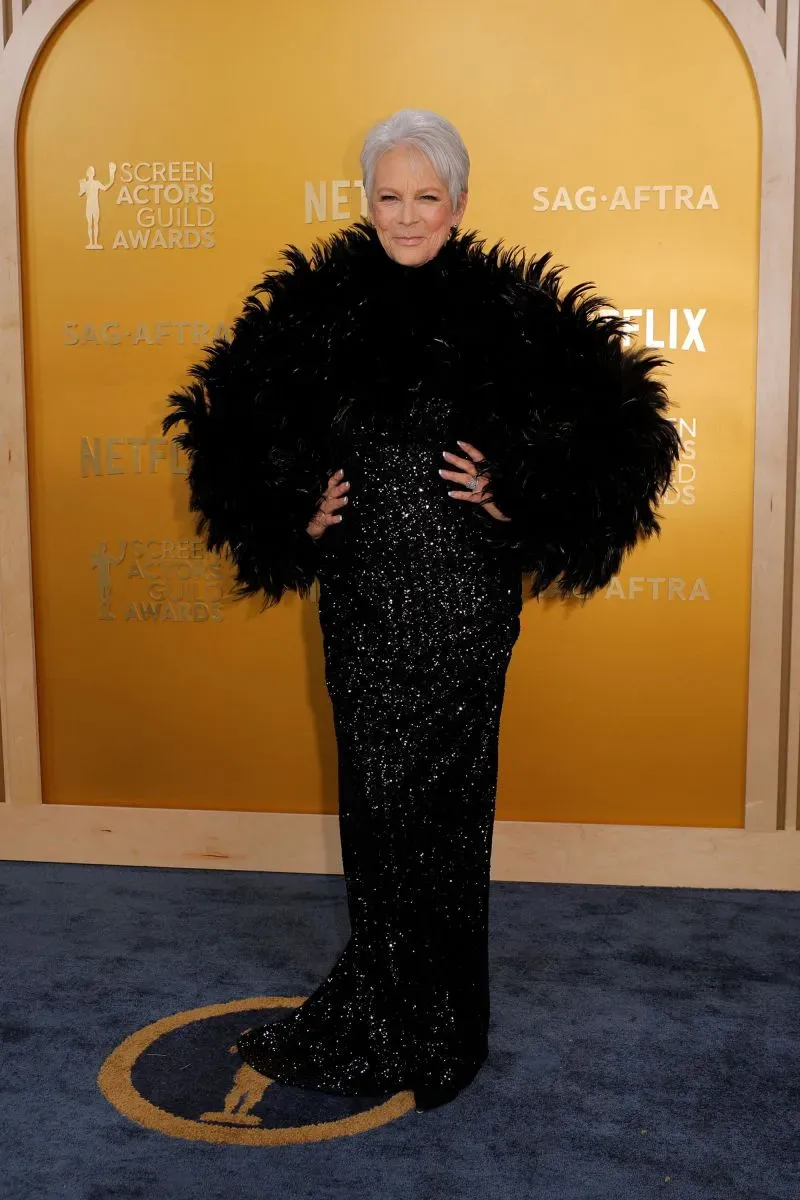 نجمة ترتدي فستاناً أسود لامعاً مع تفاصيل ريشية، تقف بفخر على السجادة الحمراء لجوائز نقابة ممثلي الشاشة، مع خلفية ذهبية.