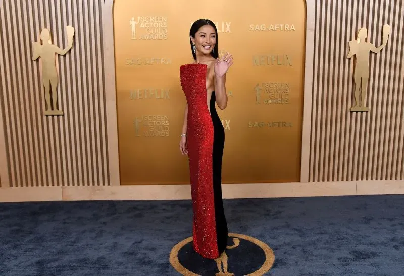 نجمة ترتدي فستاناً مميزاً يجمع بين اللونين الأحمر والأسود، تبتسم وتلوح بيدها على السجادة الحمراء خلال حفل جوائز نقابة الممثلين.