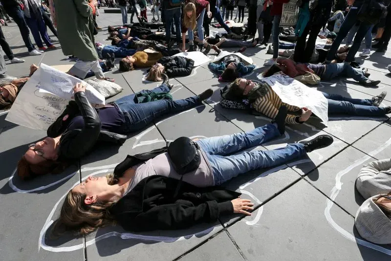 تظهر الصورة مجموعة من المتظاهرين مستلقين على الأرض في ساحة عامة، يحملون لافتات تعبر عن دعمهم لحركة #أنا_أيضًا، مما يعكس التزامهم بمناهضة العنف الجنسي.