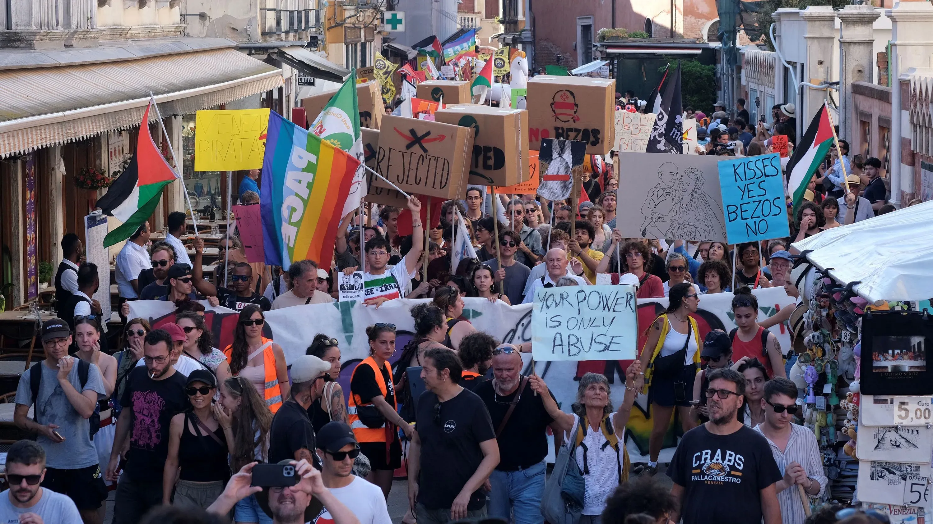تجمع حشود من المتظاهرين في شوارع البندقية، حاملين لافتات تعبر عن رفضهم لجيف بيزوس وزفافه، مع أعلام متعددة ورسائل احتجاجية.