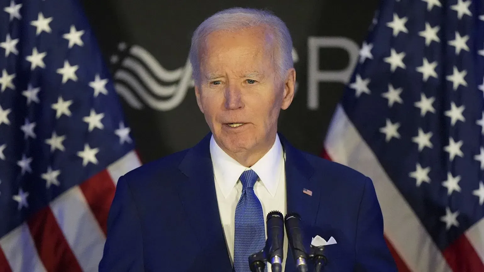 بايدن يتحدث في مؤتمر صحفي، مرتديًا بدلة زرقاء، مع العلم الأمريكي خلفه، معبرًا عن انتقاداته لترامب وسياساته المتعلقة بأوكرانيا.