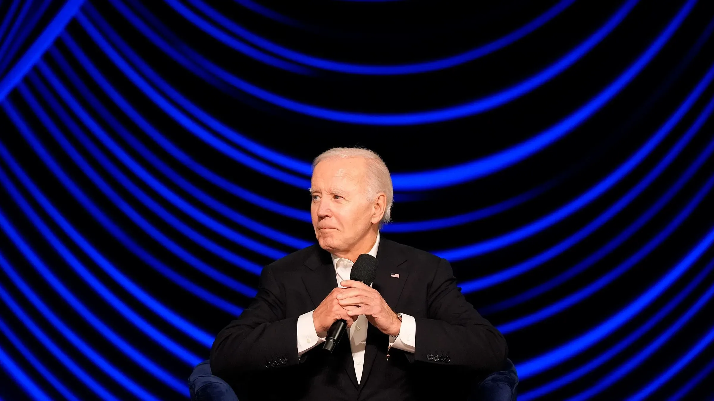 جو بايدن يجلس في حفل جمع تبرعات، يبدو متأملاً، مع خلفية زرقاء متدرجة، في إشارة إلى تدهور حالته الصحية.