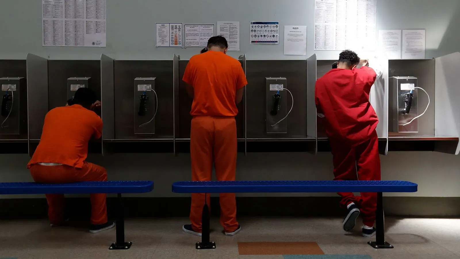 ثلاثة محتجزين يرتدون زي السجن يتحدثون عبر هواتف عمومية في منشأة احتجاز، مما يعكس ظروف احتجاز المهاجرين في السجون الخاصة.