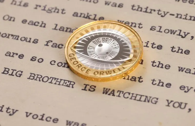 عملة تذكارية تبرز صورة عين تشبه عدسة كاميرا، تحمل نقش \"الأخ الأكبر يراقبك\" تكريمًا لجورج أورويل.