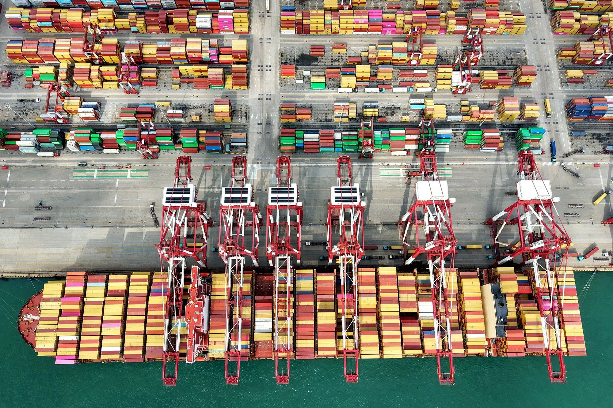 صورة جوية لميناء يحتوي على حاويات ملونة متعددة، مع رافعات تحميل كبيرة، تعكس حركة التجارة العالمية وتأثير التعريفات الجمركية.