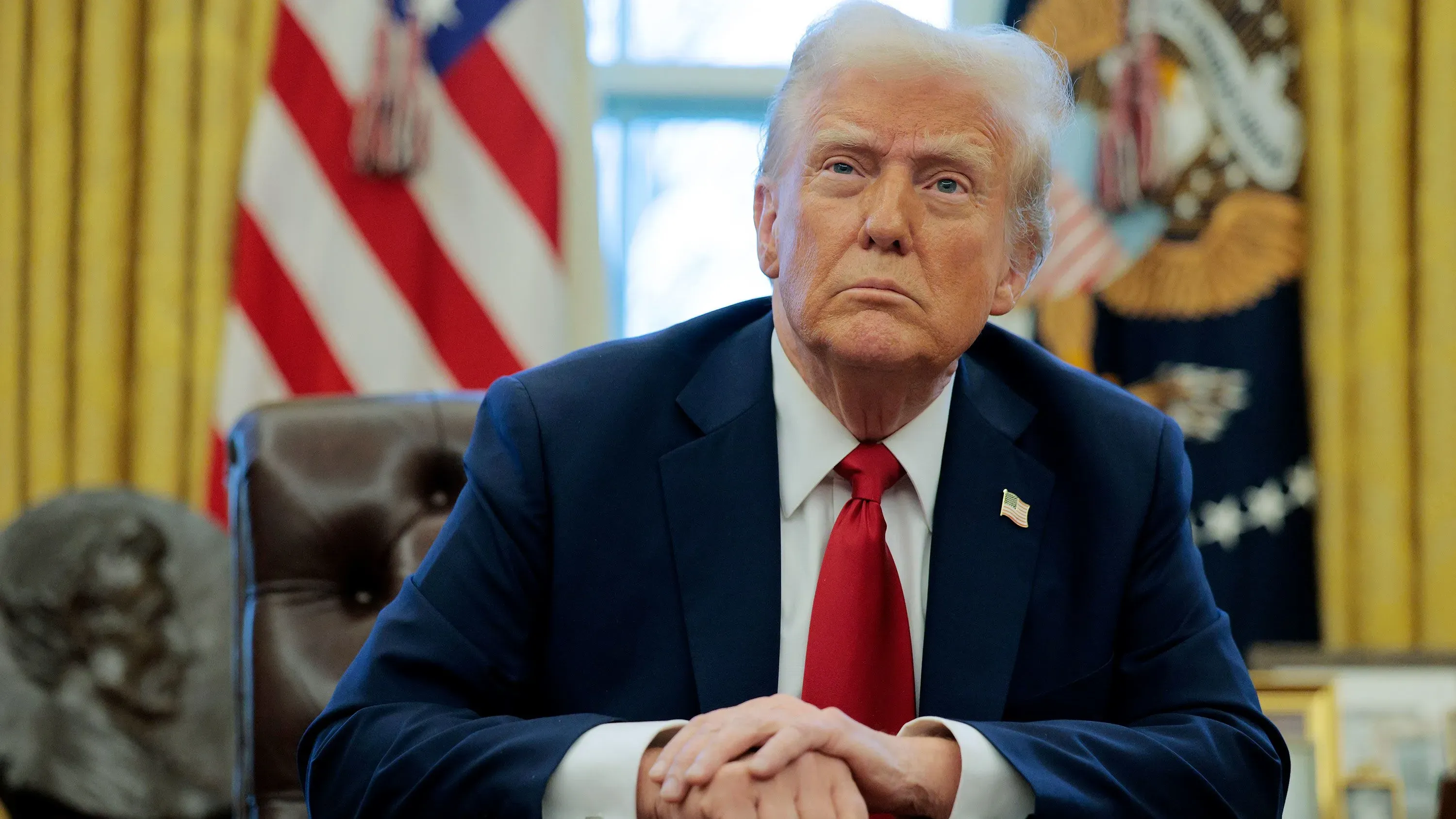 ترامب يجلس في المكتب البيضاوي، يرتدي بدلة زرقاء وقميص أبيض مع رابطة عنق حمراء، خلفه العلم الأمريكي.