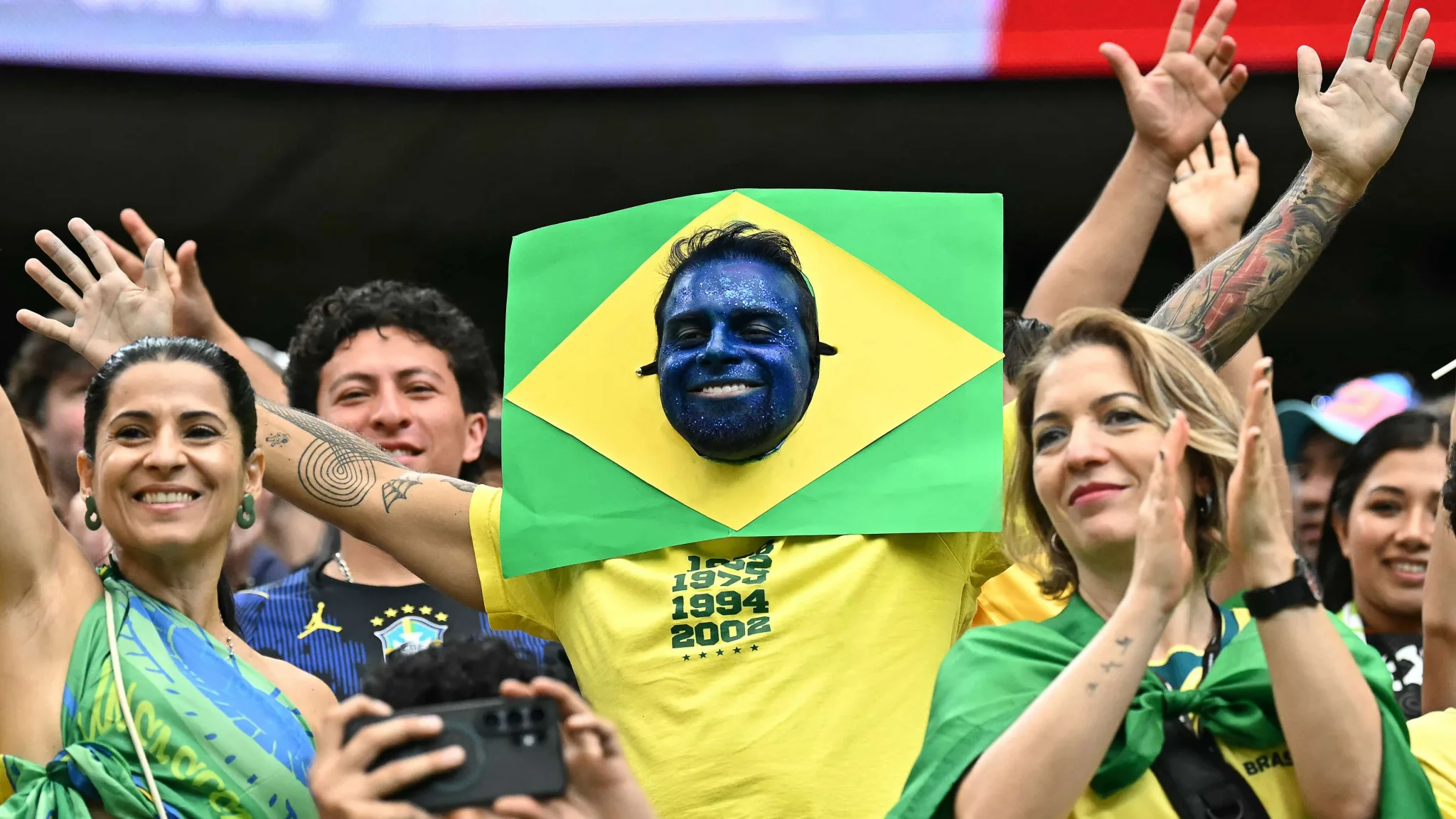 مشجع برازيلي يرفع يديه مرتدياً قناع وجه مزين بعلم البرازيل وسط حشد من المشجعين خلال كأس العالم 2026، مع التركيز على فخره بخمس ألقاب عالمية.
