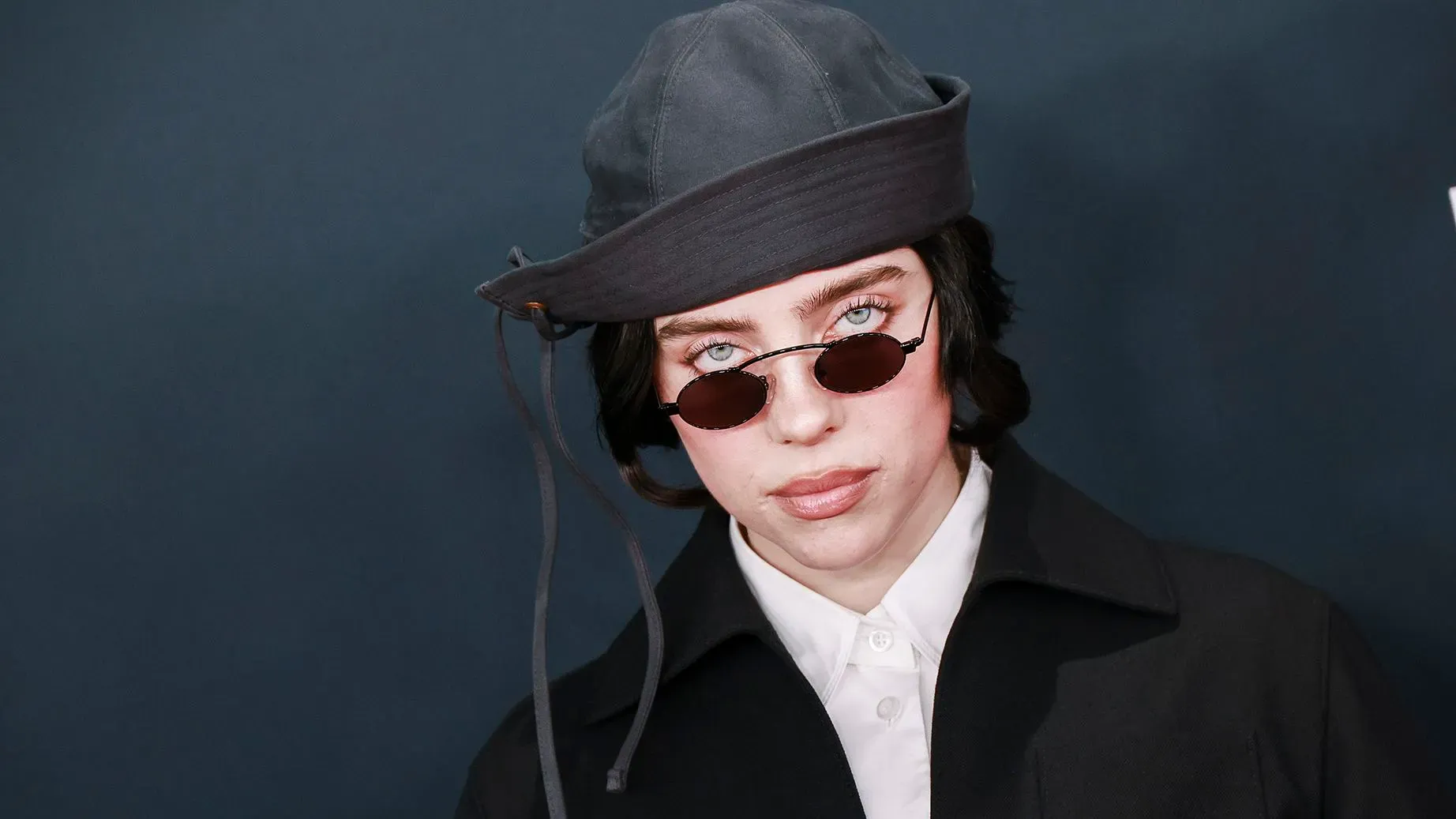 بيلي إيليش ترتدي نظارات شمسية وقبعة، تعبيرها جاد، في خلفية داكنة. تعكس الصورة أسلوبها الفريد وثقتها في نفسها.