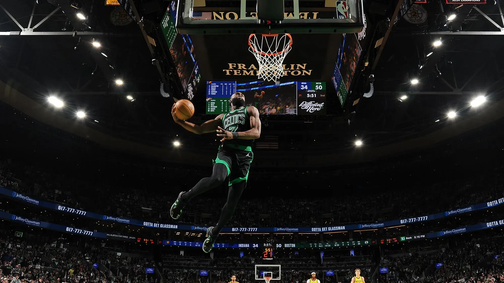 لاعب كرة سلة من فريق بوسطن سيلتيكس يقوم بعملية تسديد مثيرة في ملعب مزدحم، مع عرض للنتيجة على الشاشة الكبيرة.
