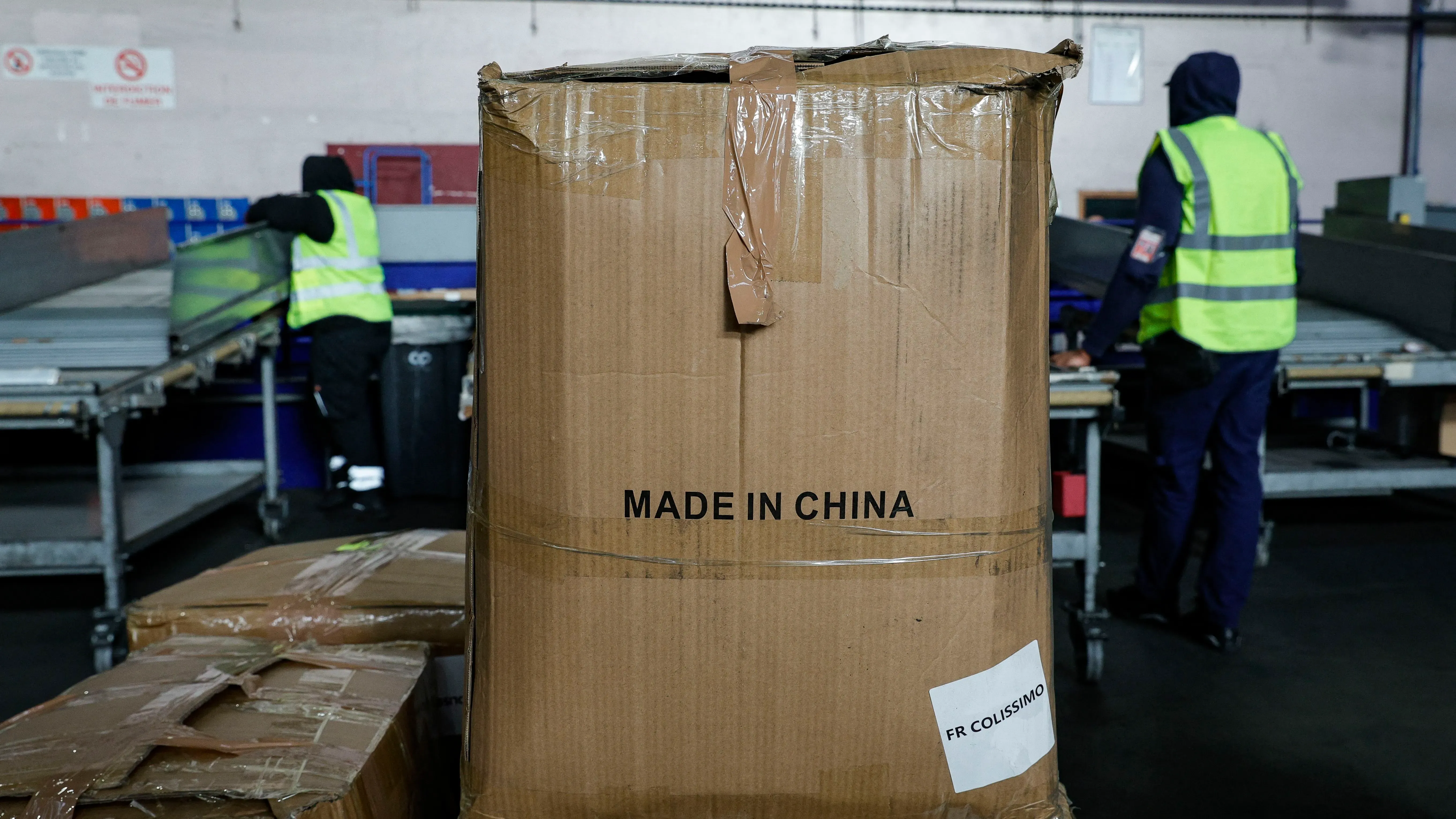 صندوق كبير مكتوب عليه "صنع في الصين" في مركز شحن، مع عمال يرتدون سترات عاكسة في الخلفية، مما يعكس التوترات التجارية بين الصين والاتحاد الأوروبي.