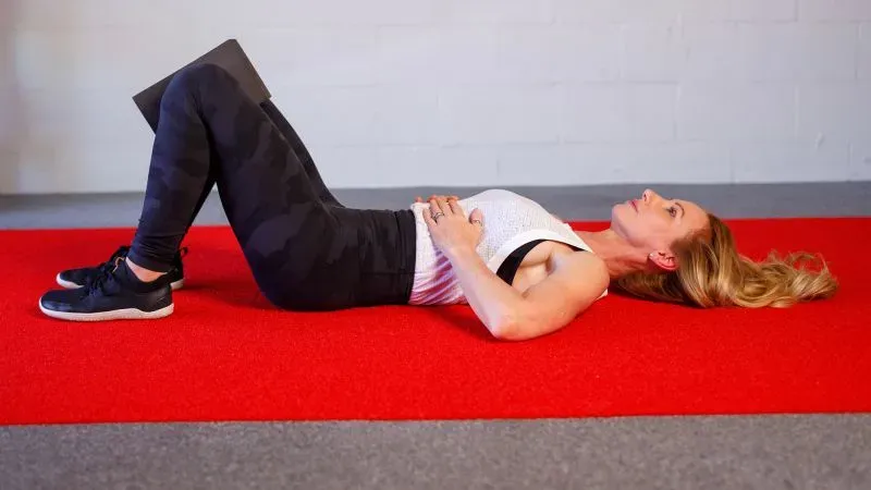 امرأة تمارس تمرين جسر التنفس المعدل على سجادة حمراء، مستلقية على ظهرها مع ثني ركبتيها، مما يعزز حركة القفص الصدري.