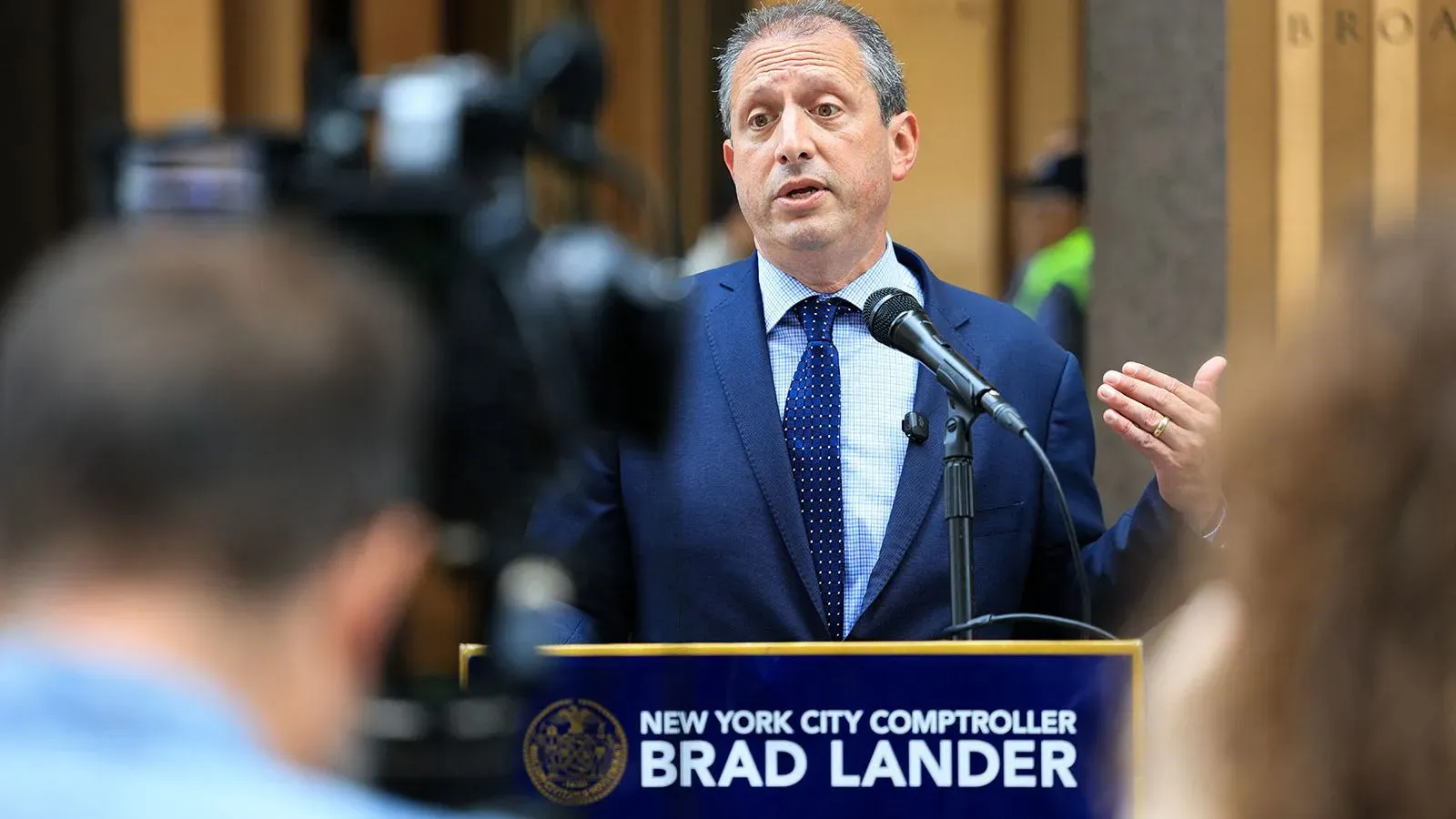 براد لاندر، المراقب المالي لمدينة نيويورك، يتحدث في مؤتمر صحفي، مع تركيز الكاميرات عليه، وسط أجواء من التوتر السياسي.