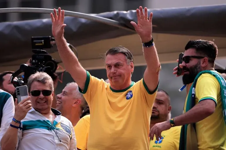 جايير بولسونارو، الرئيس البرازيلي السابق، يرفع يده أثناء تفاعله مع حشد من المؤيدين في حدث سياسي، مرتديًا قميصًا أصفر يحمل ألوان علم البرازيل.