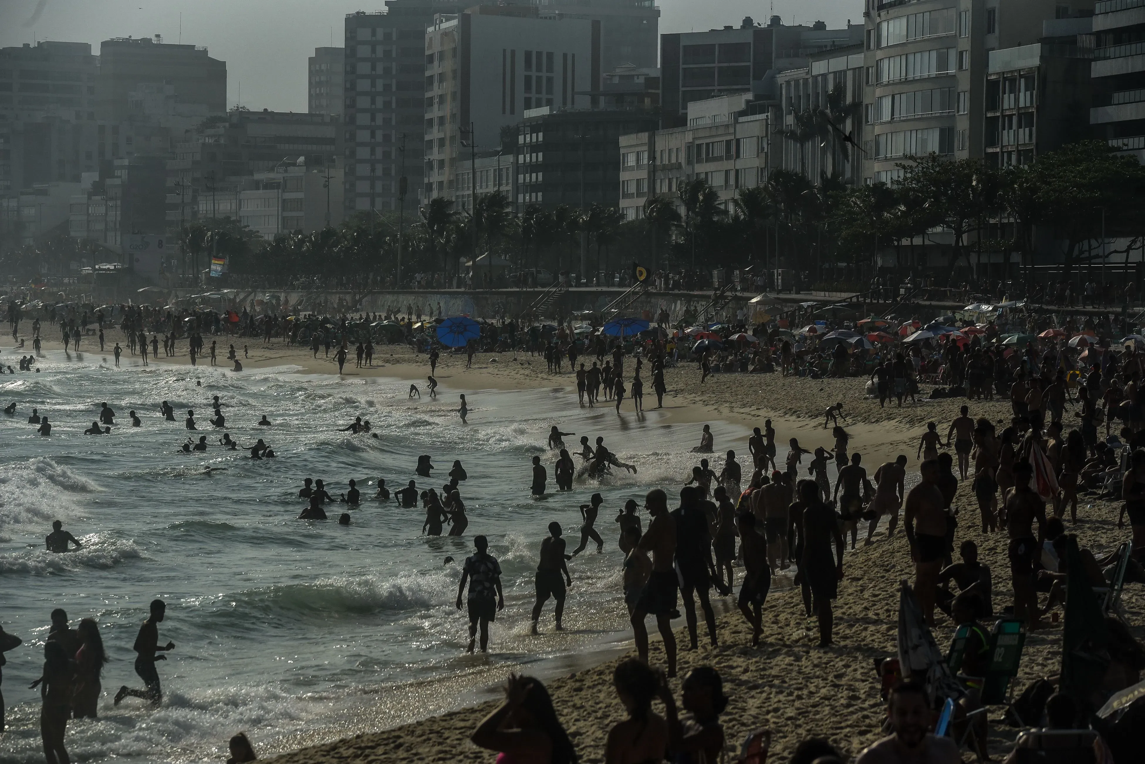 شاطئ إيبانيما في ريو دي جانيرو مزدحم بالزوار، حيث يسبح البعض في المياه بينما يستمتع الآخرون على الرمال، وسط مبانٍ شاهقة.