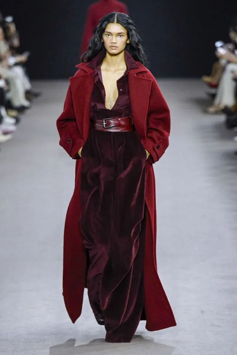 عارضة أزياء ترتدي فستاناً عميق اللون مع معطف طويل، تسير على منصة عرض الأزياء في أسبوع الموضة بميلانو، مما يعكس أسلوباً عصريًا وجريئًا.