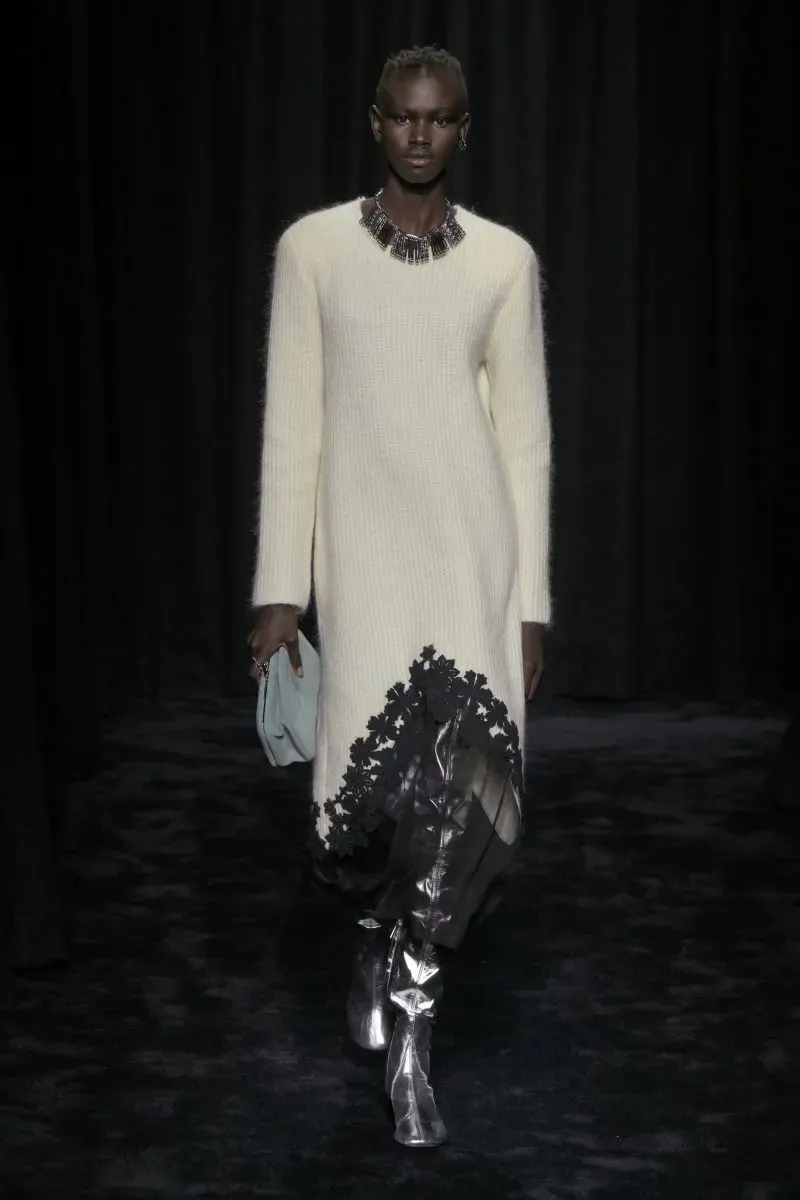 عارضة أزياء ترتدي فستانًا أبيض طويلًا مزينًا بحواف سوداء، مع إكسسوارات فضية، تسير على منصة عرض مظلمة.