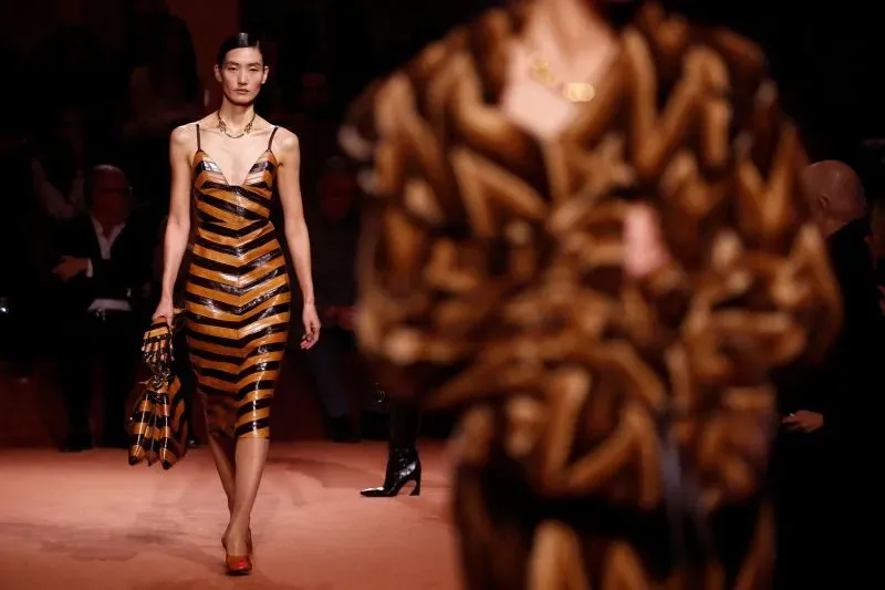 عارضة أزياء ترتدي فستانًا مخططًا بالأصفر والأسود، تحمل حقيبة متناسقة، بينما تتقدم على منصة عرض أزياء في ميلانو.