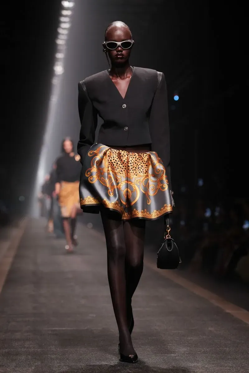 عارضة أزياء ترتدي تصميمًا عصريًا يجمع بين سترة داكنة وتنورة مزينة بنقوش ذهبية، تسير على مدرج أسبوع الموضة في ميلانو.