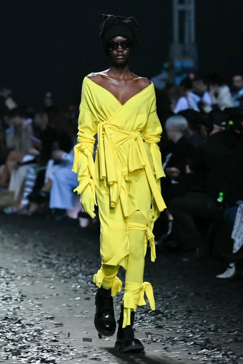 عارضة أزياء ترتدي فستانًا أصفر لامعًا بأكمام متدلية، مع نظارات شمسية وقبعة سوداء، تسير على منصة عرض مليئة بالقصاصات.