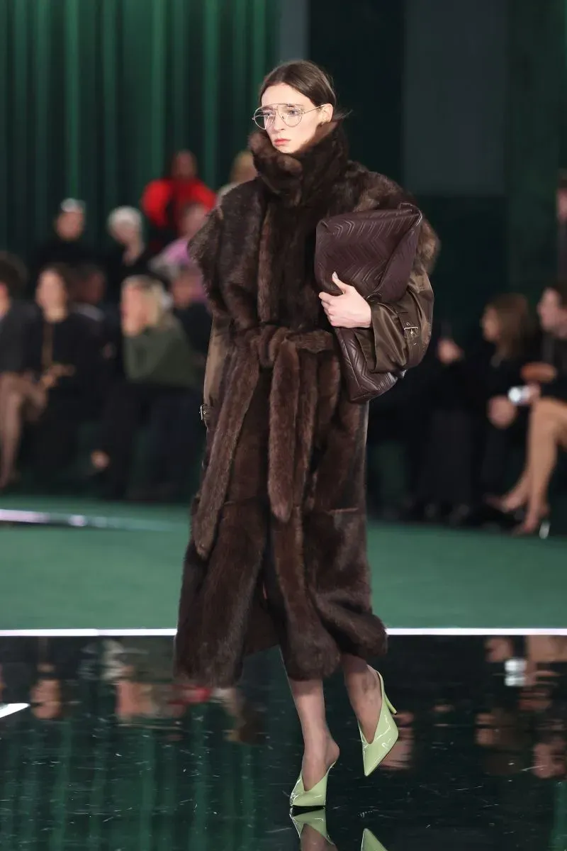 عارضة أزياء ترتدي معطفًا بنيًا من الفرو مع نظارات كبيرة وحقيبة، تسير على منصة عرض أزياء في ميلانو.