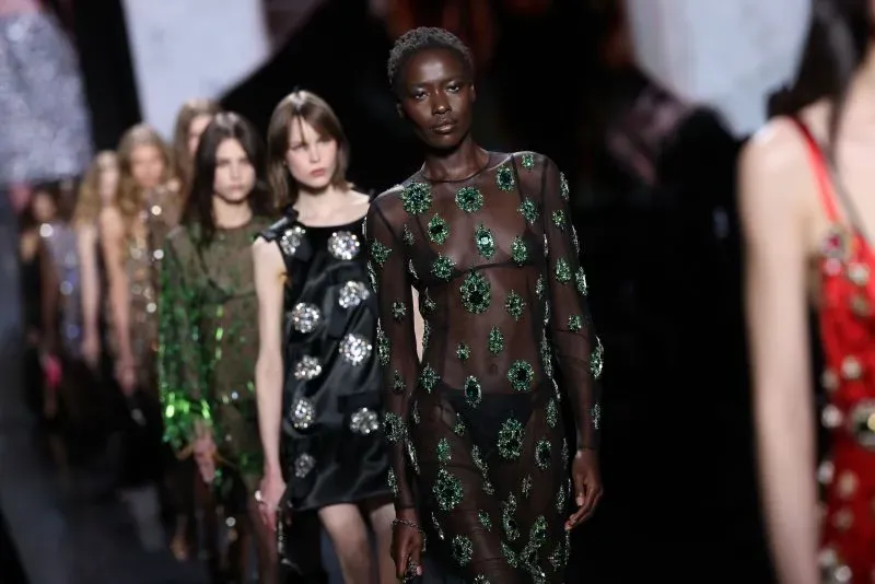 عارضة أزياء ترتدي فستانًا شفافًا مزينًا بأحجار كريمة خضراء، تتقدم على المنصة بينما تسير عارضات أخريات خلفها في عرض أزياء ميلانو.