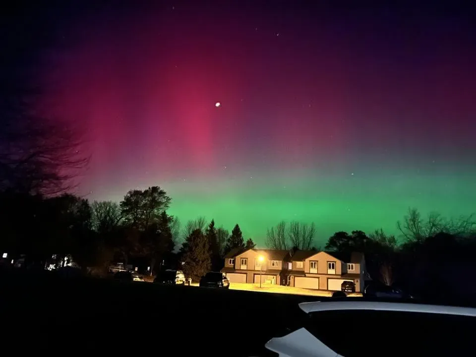شفق قطبي ملون يظهر في سماء الليل، مع أضواء خضراء وحمراء تتراقص فوق المنازل، نتيجة لعاصفة مغناطيسية G4.