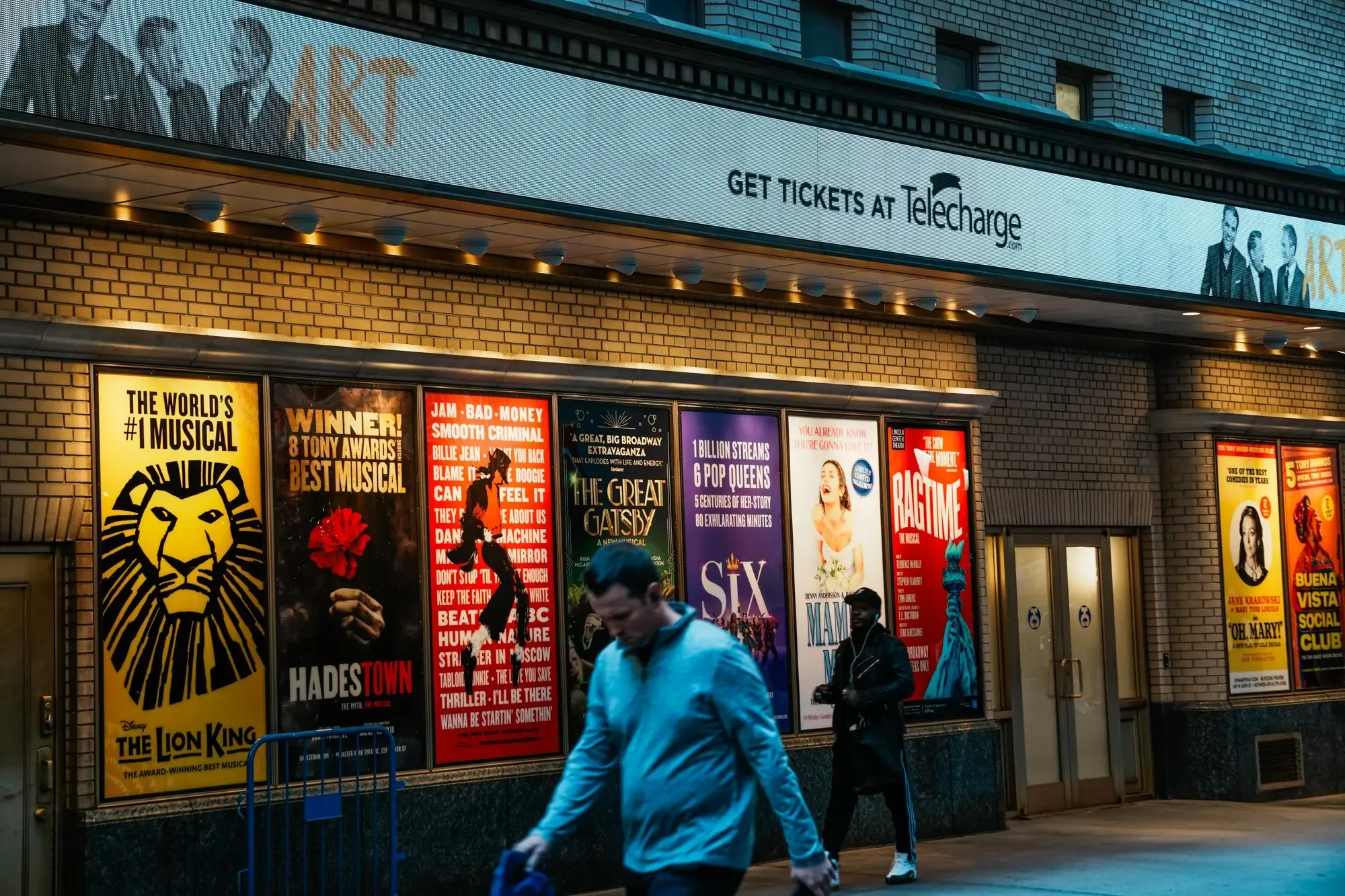 واجهة مسرح برودواي تعرض ملصقات لمسرحيات موسيقية شهيرة، مع أشخاص يتجولون في المنطقة، مما يعكس حيوية الثقافة الفنية في نيويورك.