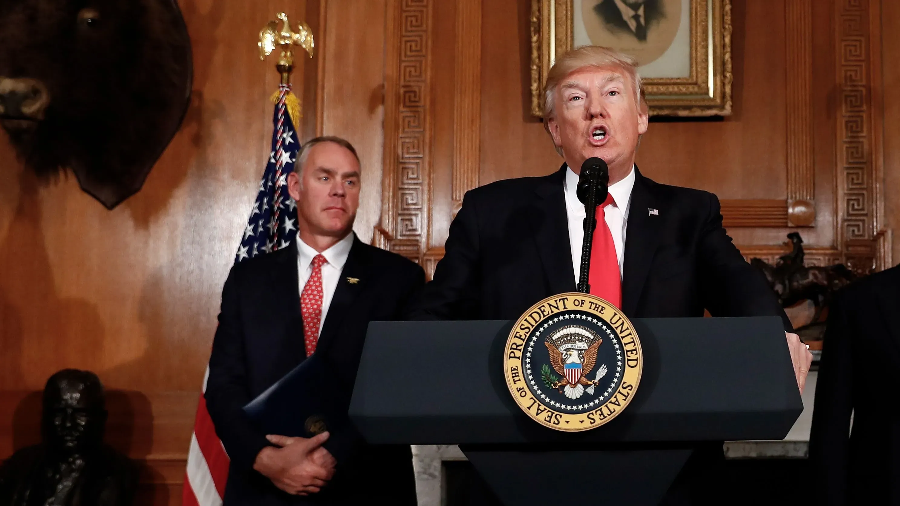 النائب ريان زينكي يتحدث أمام الميكروفون خلال مؤتمر صحفي، مع العلم الأمريكي خلفه وصور حيوانات على الجدران، معربًا عن مخاوفه بشأن الميزانية.