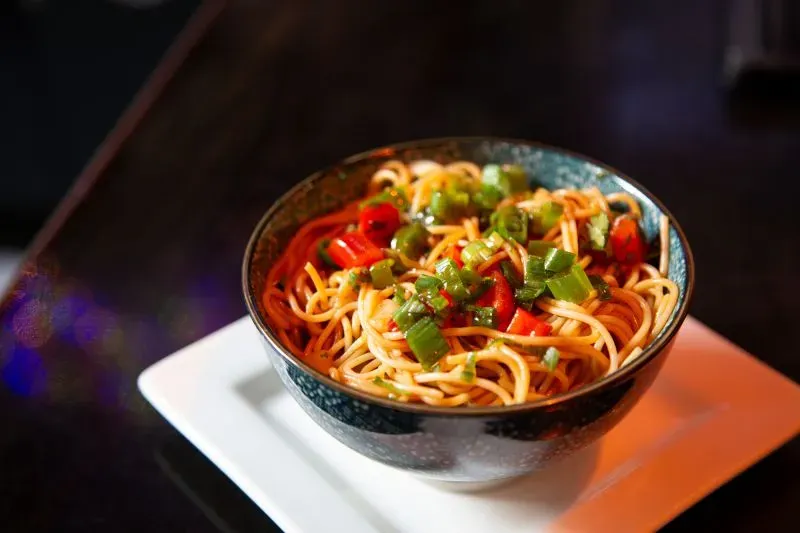 وعاء من المعكرونة الملونة مع قطع من الفلفل الأحمر والفلفل الأخضر، يقدم على طبق أبيض، يعكس تنوع المأكولات في بافالو.