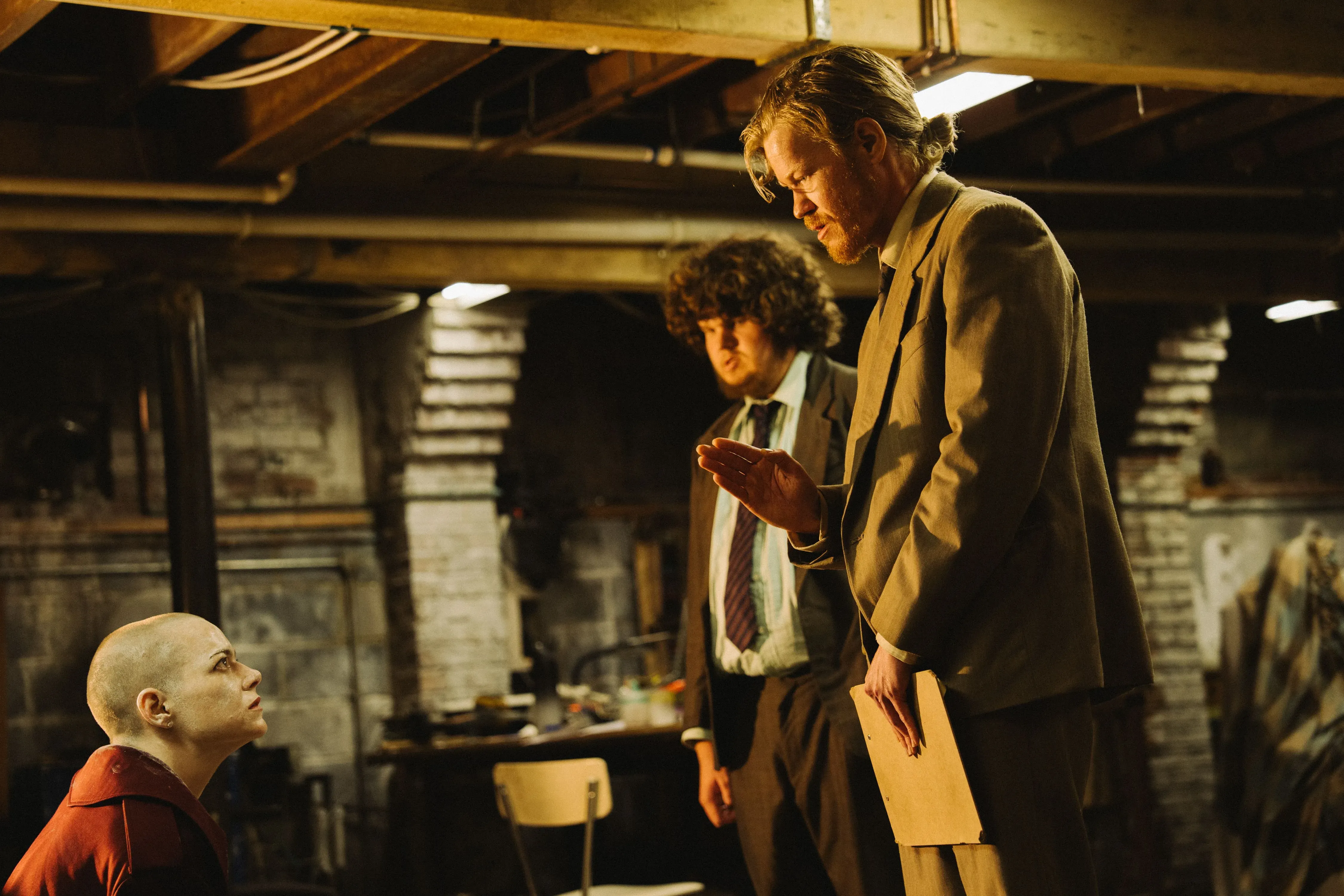 مشهد من فيلم "بوغونيا" يظهر شخصية ميشيل ذات الرأس الحليق محاطة بشخصين في قبو، حيث تتصاعد التوترات حول مؤامرات الكائنات الفضائية.