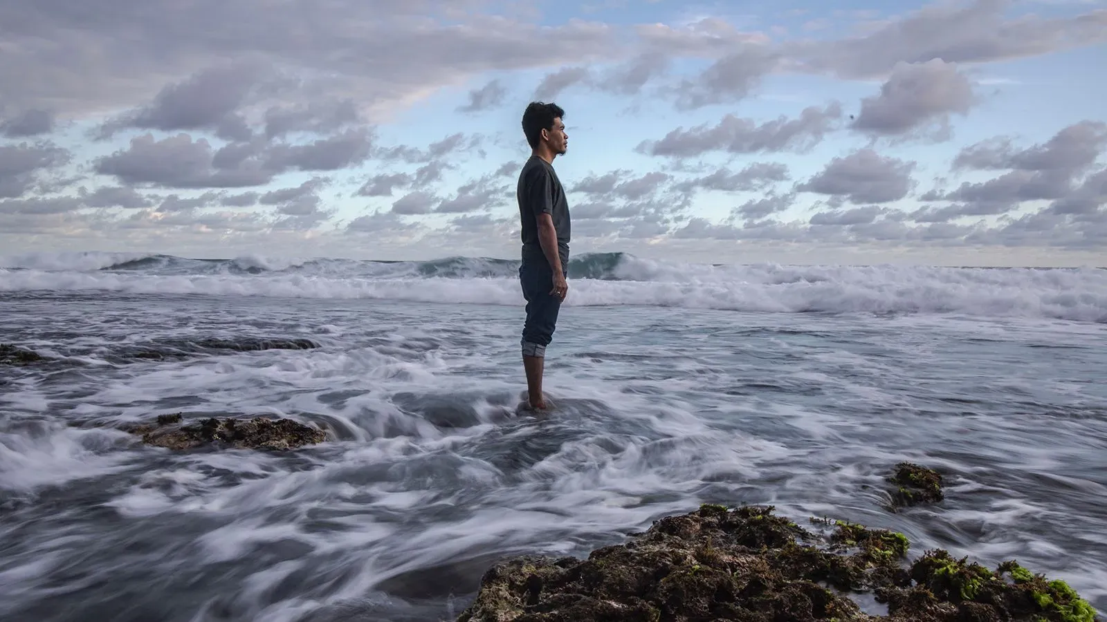 رجل يقف في الماء مع الأمواج تتلاطم حوله، يعكس مشاعر الألم والمعاناة التي واجهها أثناء عمله في صناعة صيد الأسماك.