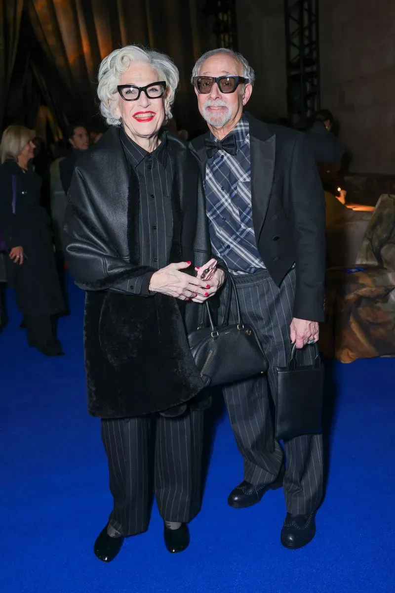 زوجان مسنّان يرتديان ملابس أنيقة باللونين الأسود والرمادي، يقفان معًا في حدث رسمي على سجادة زرقاء.