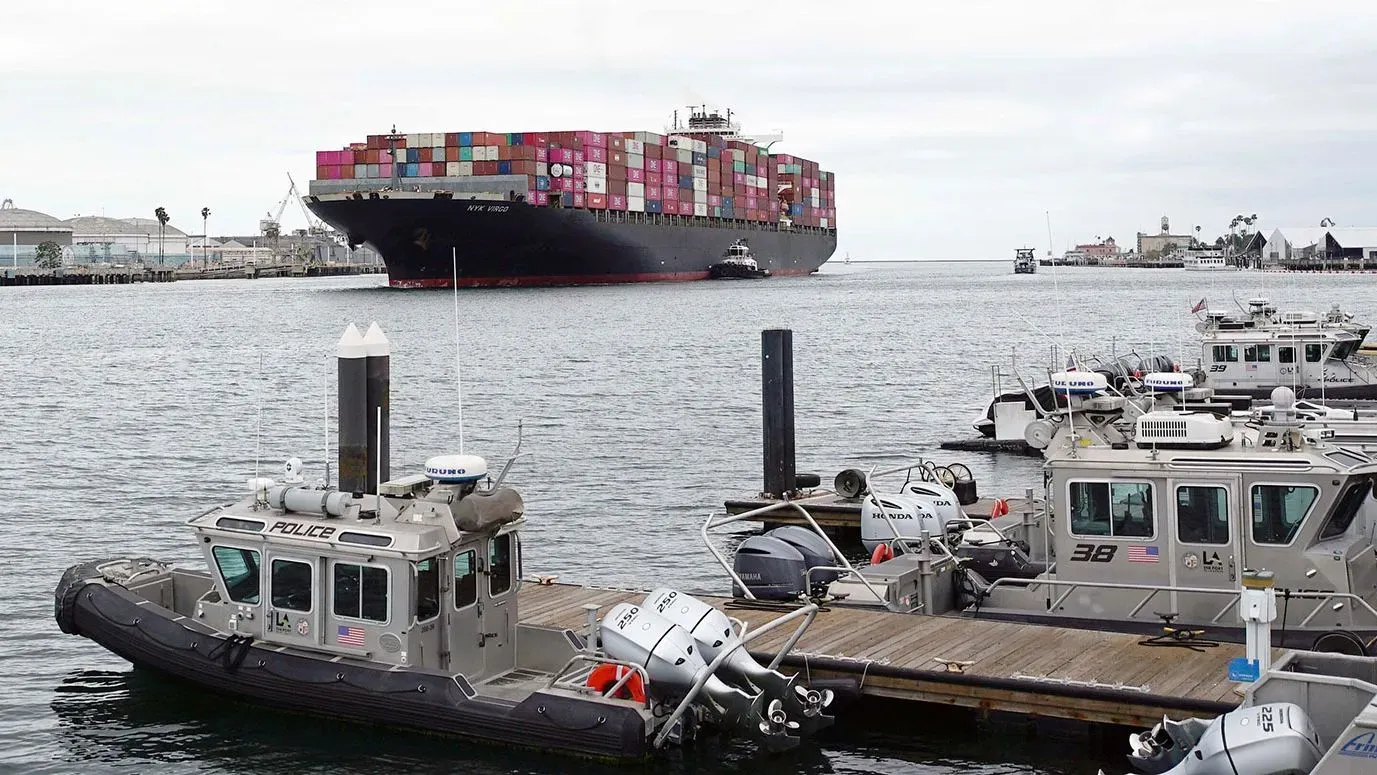 سفينة شحن كبيرة محملة بالحاويات الملونة تبحر في الميناء، مع قوارب شرطة صغيرة راسية على الرصيف، تعكس حركة التجارة الدولية.