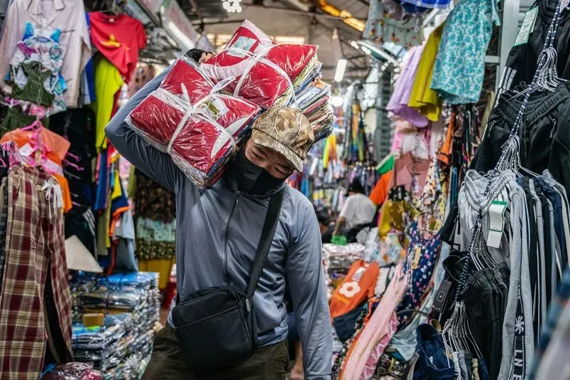 عامل يحمل حزمًا من الملابس في سوق مزدحم، يعكس تأثير التعريفات الجمركية على صناعة الملابس في كمبوديا ودول جنوب شرق آسيا.