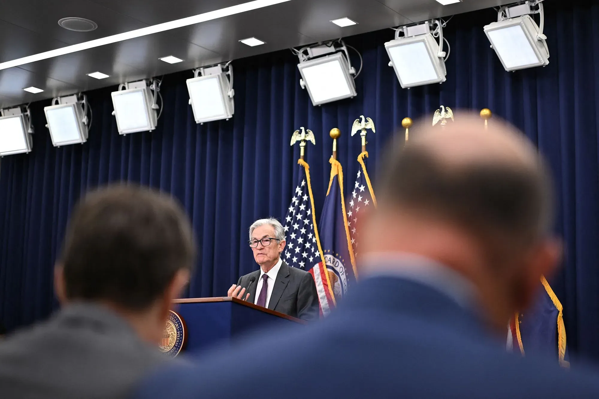 رجل مسن يتحدث في مؤتمر صحفي، مع أعلام أمريكية خلفه وأضواء ساطعة، يعكس تراجع سوق الإسكان في الولايات المتحدة.