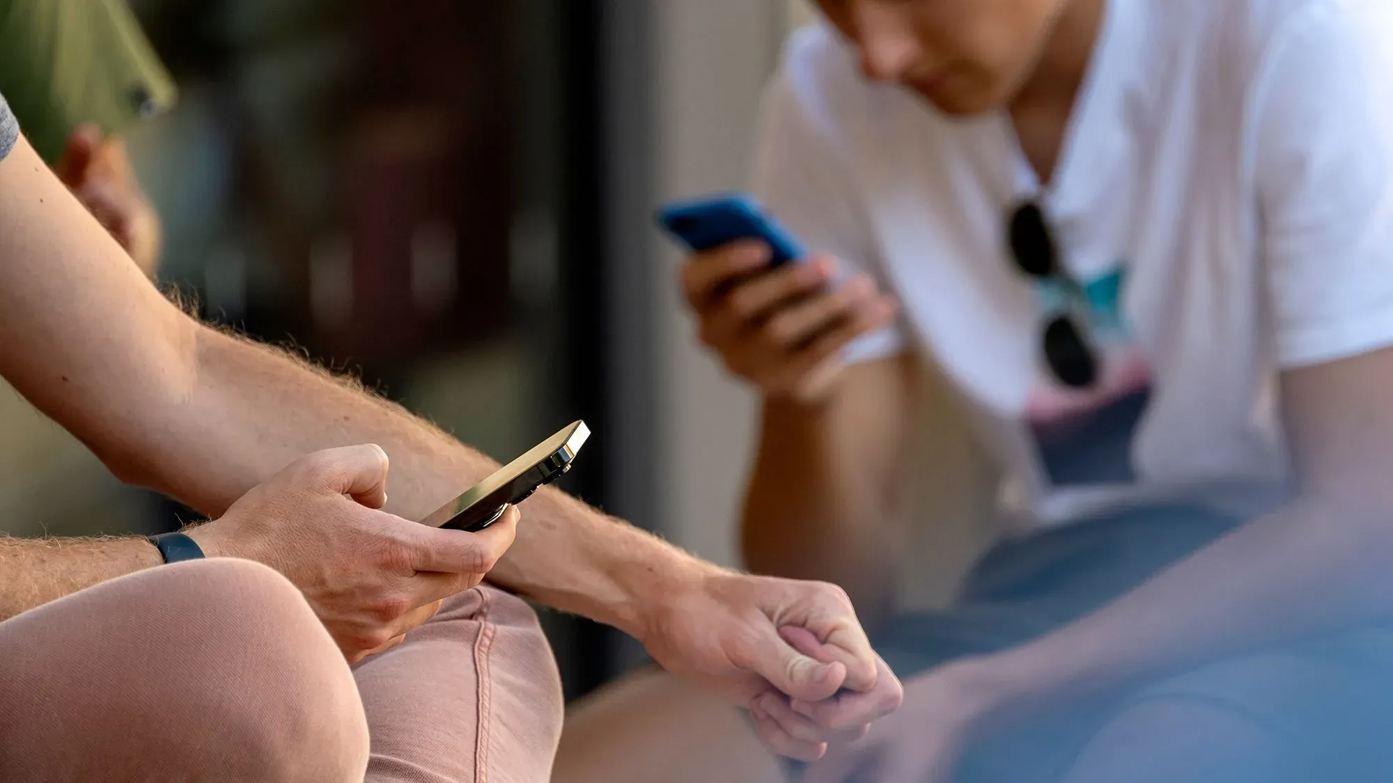 شخصان يجلسان مع هواتف ذكية، أحدهما يستخدم هاتفاً بيديه بينما الآخر يتصفح هاتفه. يُظهر المشهد تزايد شراء الهواتف المستعملة.