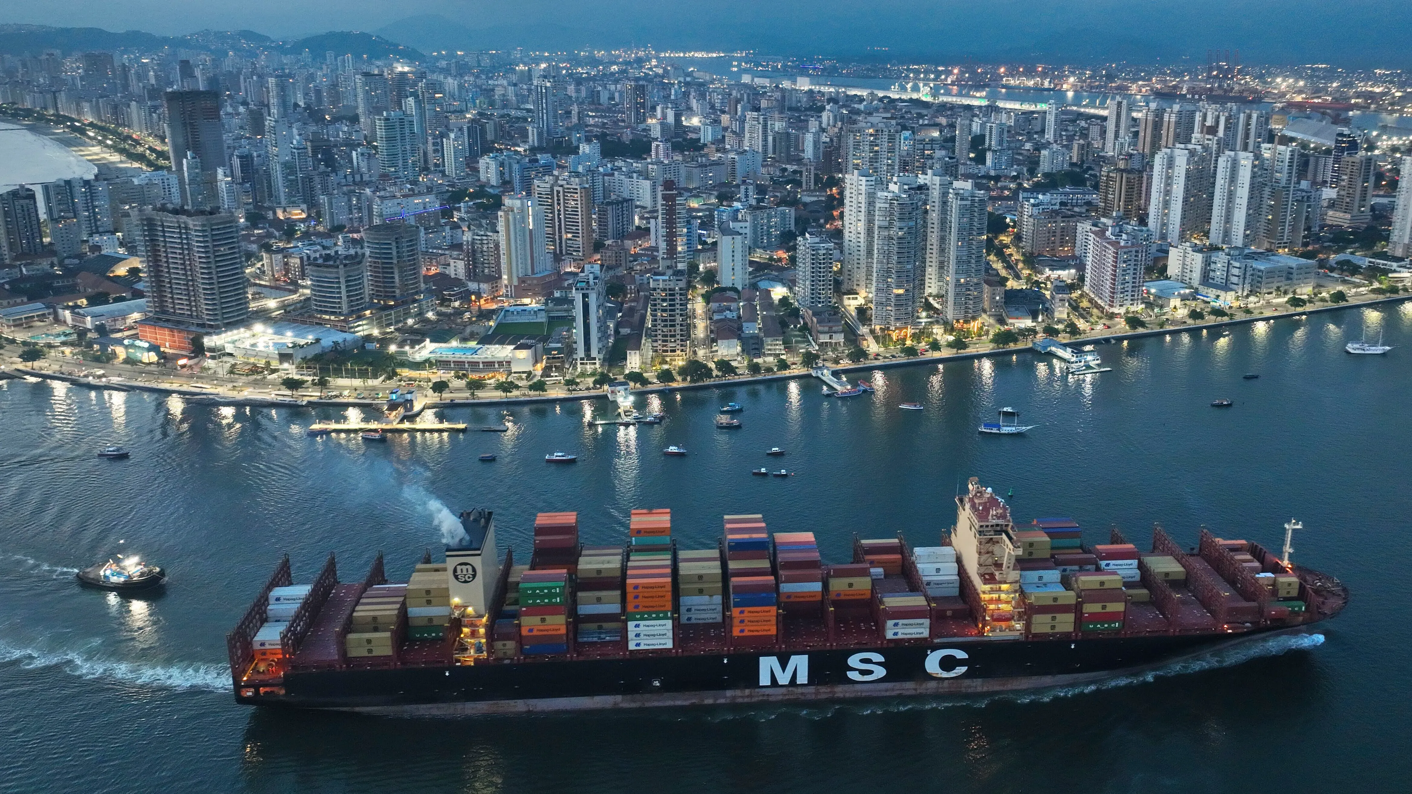 سفينة شحن تحمل حاويات ملونة تبحر في نهر بجوار مدينة حديثة، مما يعكس تأثير التعريفات الجمركية على التجارة العالمية.