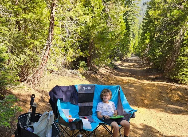 طفل يجلس على كرسي تخييم أزرق في غابة، محاط بأشجار كثيفة، بينما يبدو الطريق الترابي خلفه خاليًا.