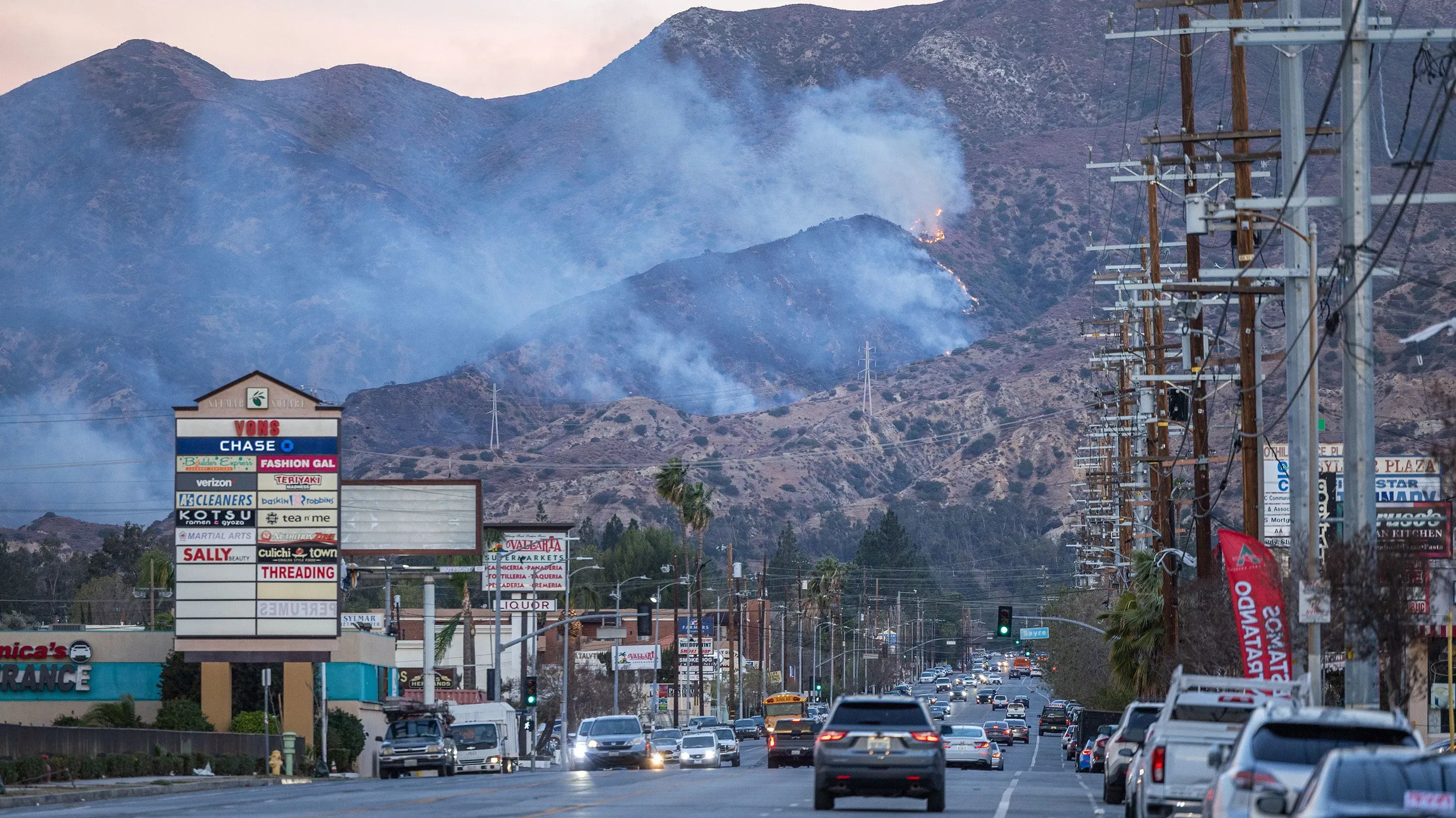 تصاعد الدخان من حريق في الجبال بالقرب من لوس أنجلوس، مع وجود سيارات على الطريق وواجهة متاجر في المقدمة.