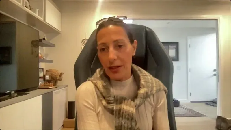 امرأة تتحدث في مقابلة، ترتدي سكارفًا وتجلس أمام شاشة تلفاز، تعبر عن مشاعرها حول حرائق الغابات وتأثيرها على المجتمع.