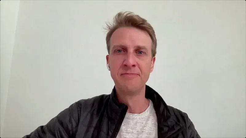 صورة لرجل مبتسم يرتدي سترة داكنة وقميص رمادي، أمام خلفية بيضاء، تعكس مشاعر الأمل وسط الظروف الصعبة.